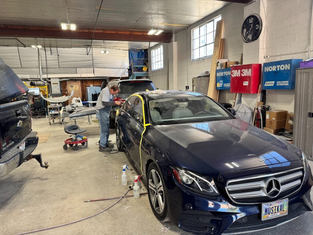Auto body technician repairing a dark blue Mercedes sedan inside a professional collision repair shop, with tools, equipment, and other vehicles in the background.